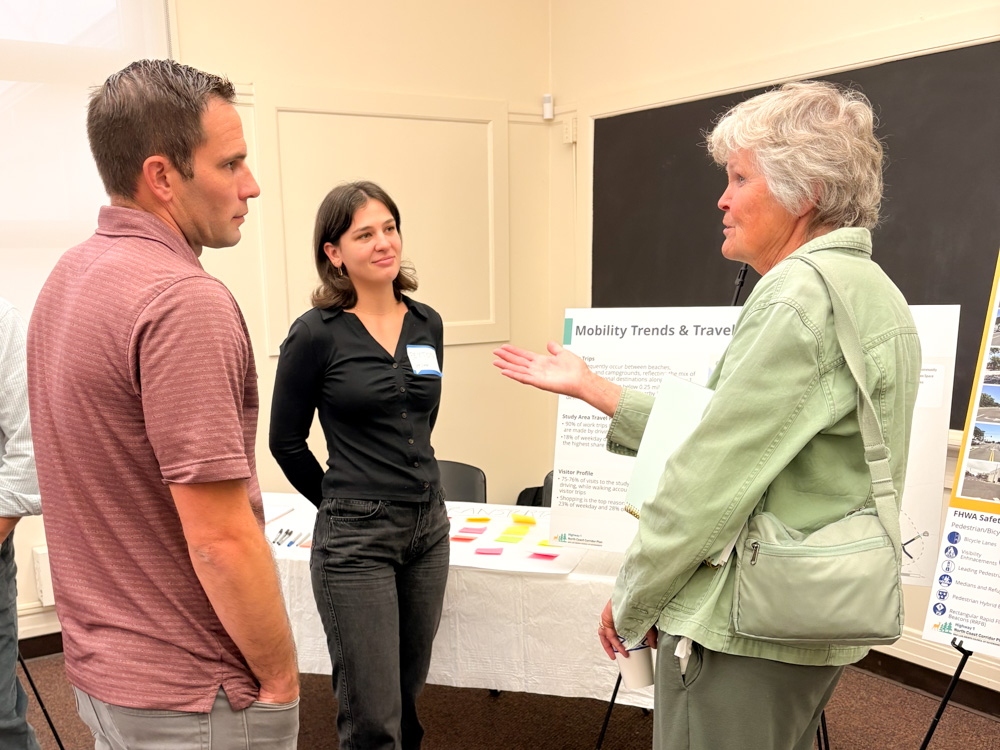 A group of community members and staff gather inside a brightly lit room at the Coast Unified School District for a Visioning Workshop, chatting in small groups and viewing information boards set up on easels.